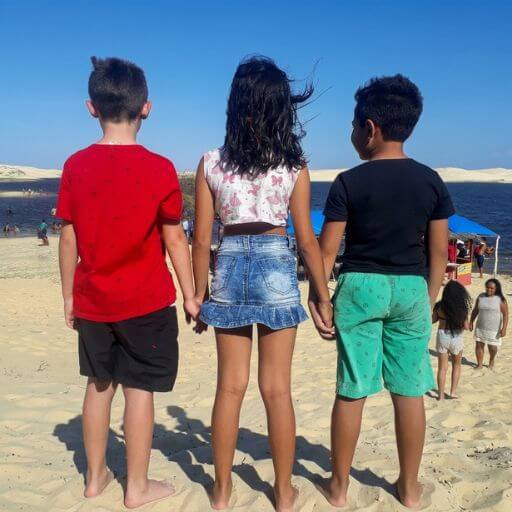Três crianças de mãos dadas de costas a olhar para o mar numa praia ensolarada, representando a jornada de crescimento e a orientação familiar.