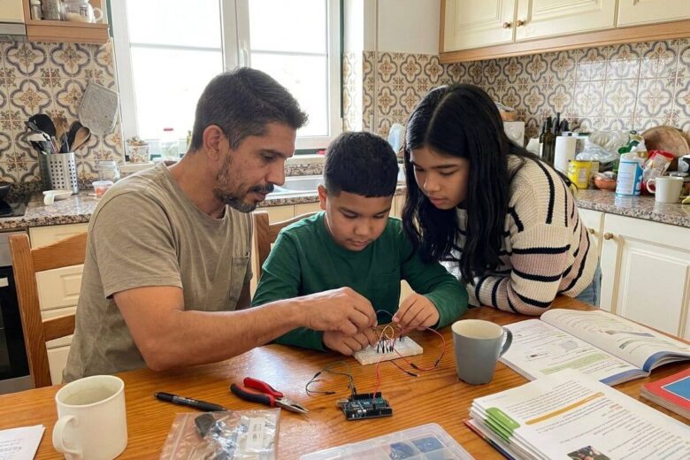 Fotografia espontânea e real de um pai [P1] sentado à mesa da cozinha, ensinando pacientemente o seu filho adolescente [P3] e a sua filha mais nova [P4] a montarem um pequeno projeto de madeira.
